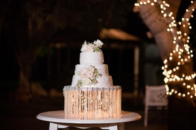 A beautifully decorated wedding cake stands tall against a backdrop of twinkling lights. The cake is multi-tiered and adorned with delicate sugar flowers and intricate piping. The ambiance is romantic and celebratory.